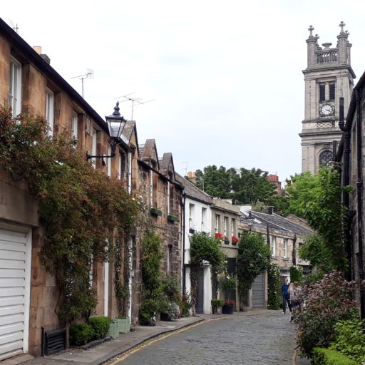 narrow residential road with greenery on curved terraced buildings and a church tower visible at the end of the street